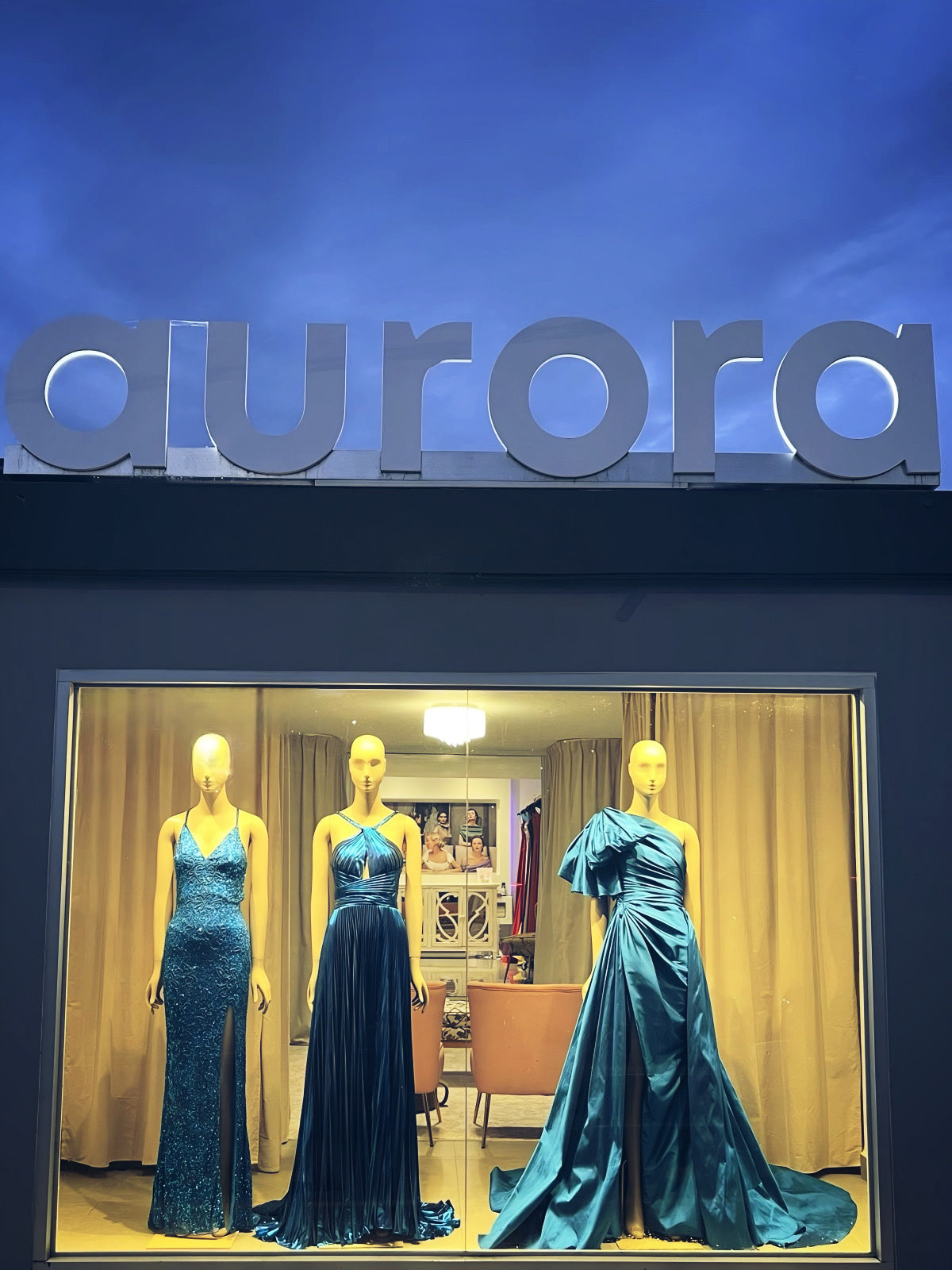 Display of three mannequins wearing blue evening gowns in a store window with 'aurora' branding above.